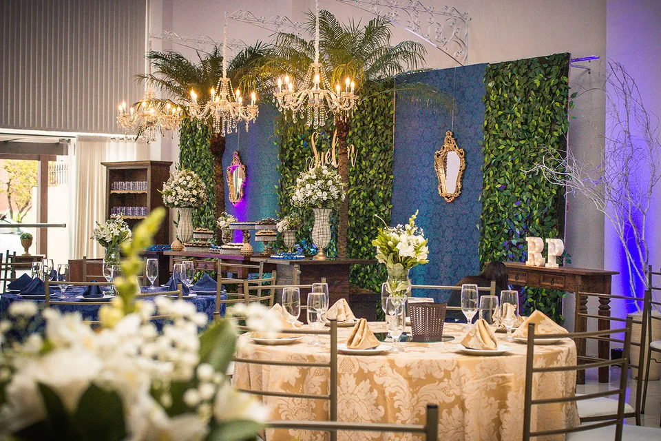 Mesa de convidados com toalha dourada, arranjos de flores brancas e lustres de cristal